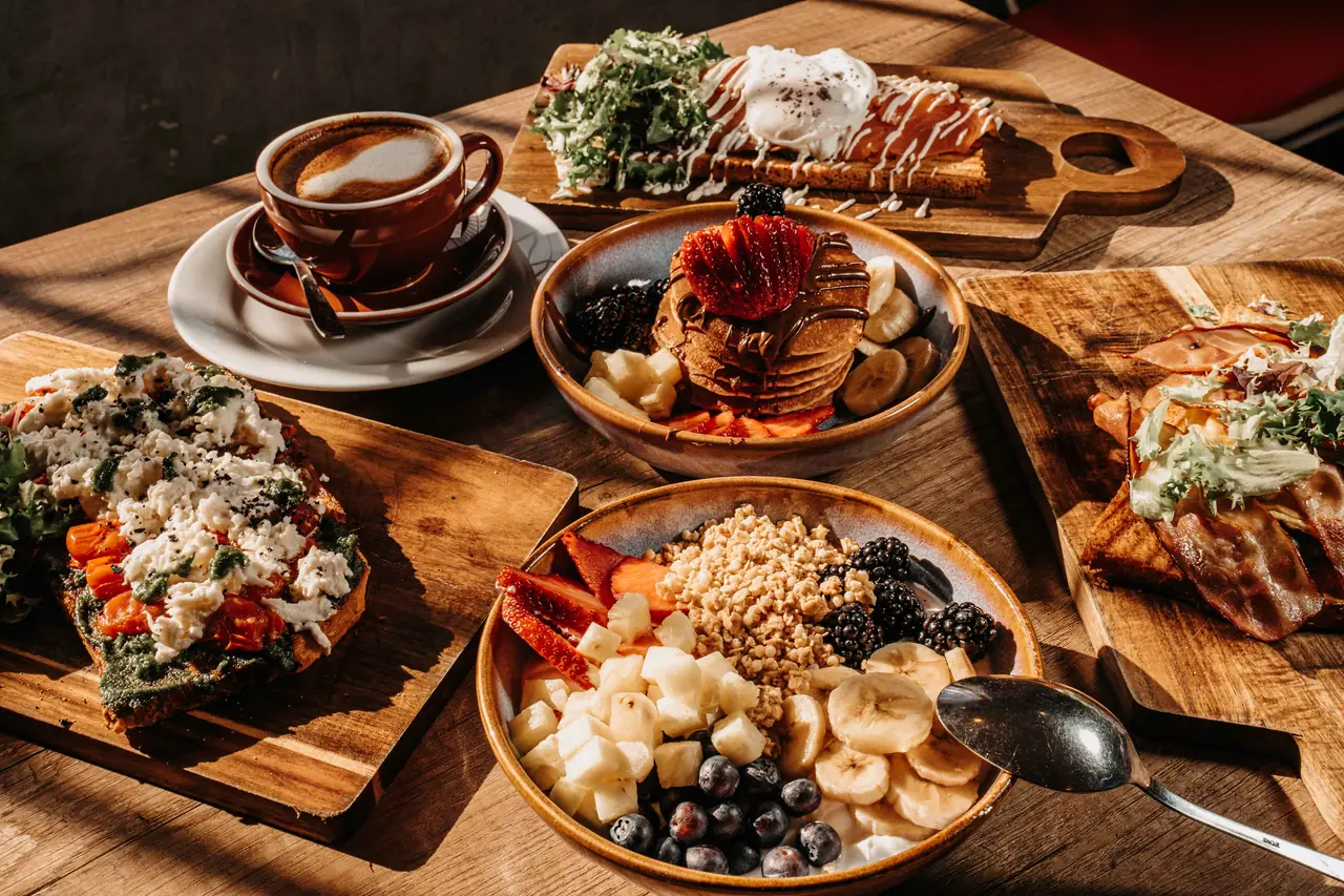 Mesa de desayuno con tostadas, bowl de fruta, tortitas y café bañada por el sol